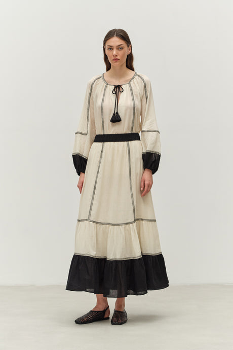 Off-white long-sleeve maxi dress with black trim, tassel neckline, and gathered cuffs.