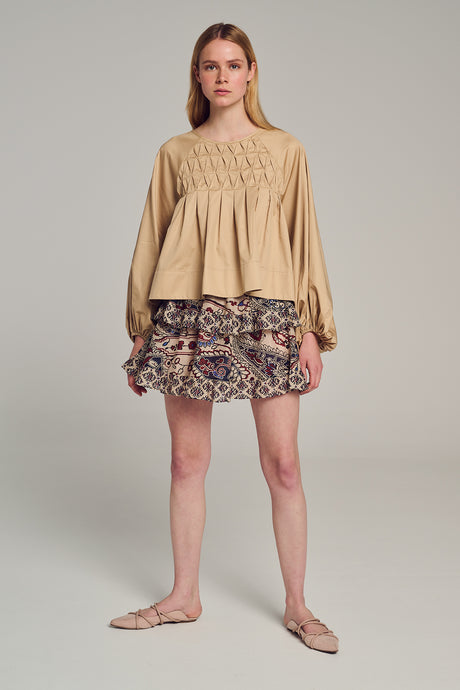 Beige long-sleeve blouse with smocked front and gathered waist, styled with patterned mini skirt.