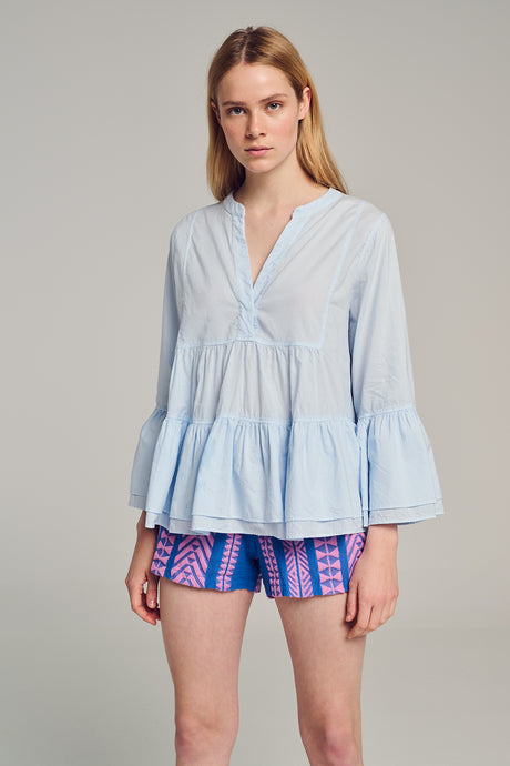 Light blue long-sleeve blouse with V-neckline and tiered ruffle hem worn with colorful shorts.