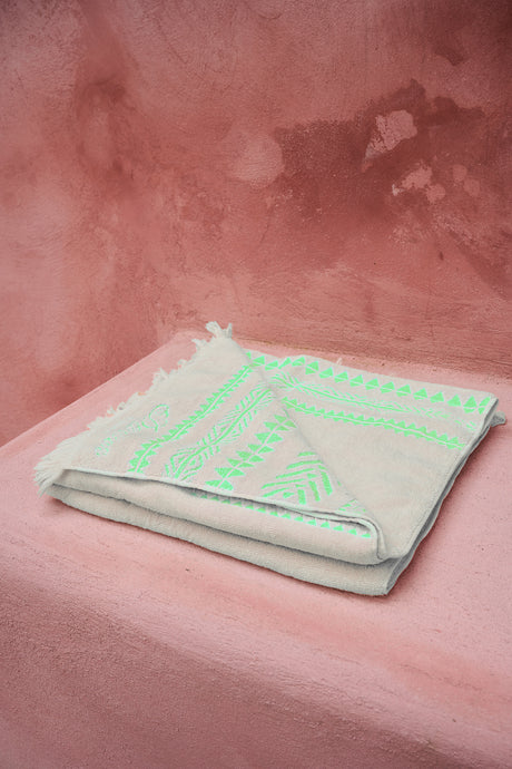 Folded offwhite beach towel with green geometric patterns and fringed edges on pink surface.