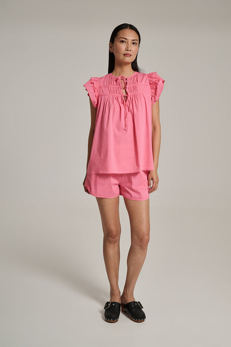 Pink popeline short-sleeve top with ruffled shoulders and matching shorts, styled with black slip-on shoes.