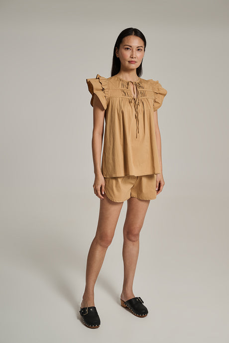 Beige sleeveless blouse with ruffled shoulders and tie neckline, paired with matching shorts and black slip-on shoes.