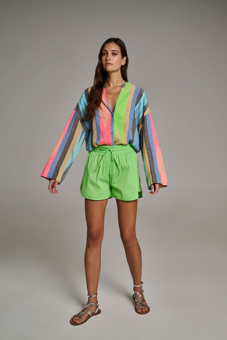 Striped long-sleeve blouse in pastel colors paired with bright green high-waist shorts and flat metallic sandals.
